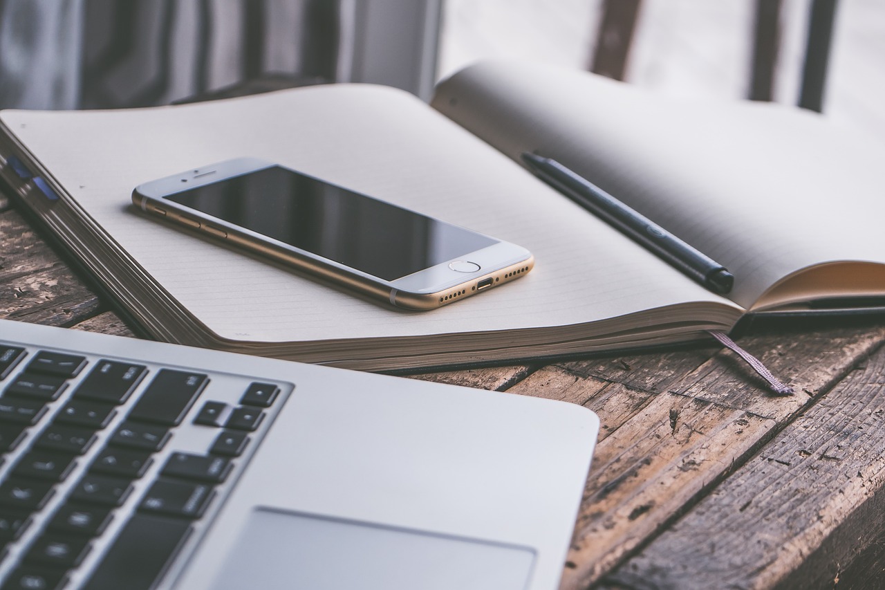 Ein offenes Notizbuch, ein Smartphone und ein Laptop auf einem Holztisch