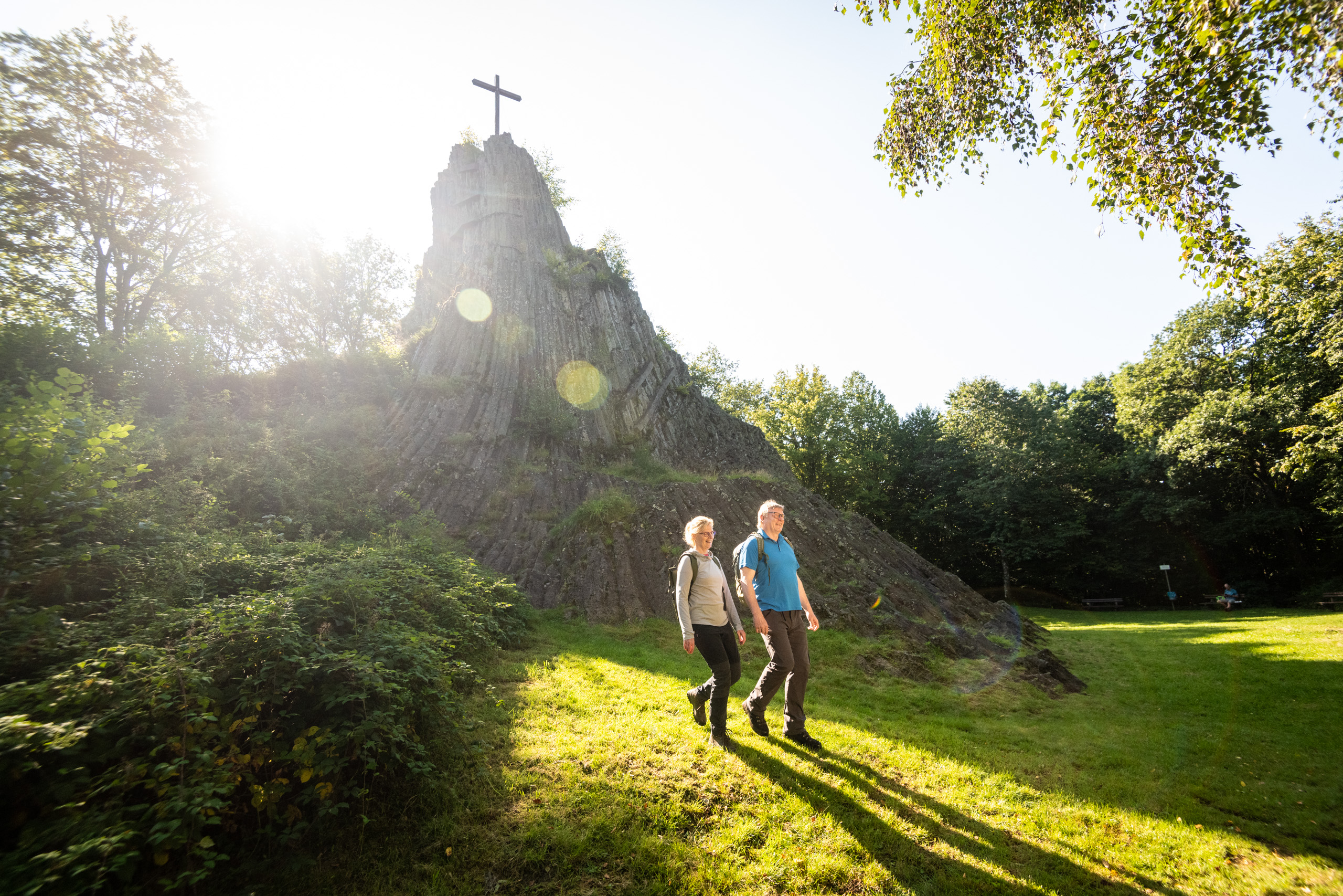 Zwei Personen spazieren neben einem Felsen mit einem Kreuz auf der Spitze