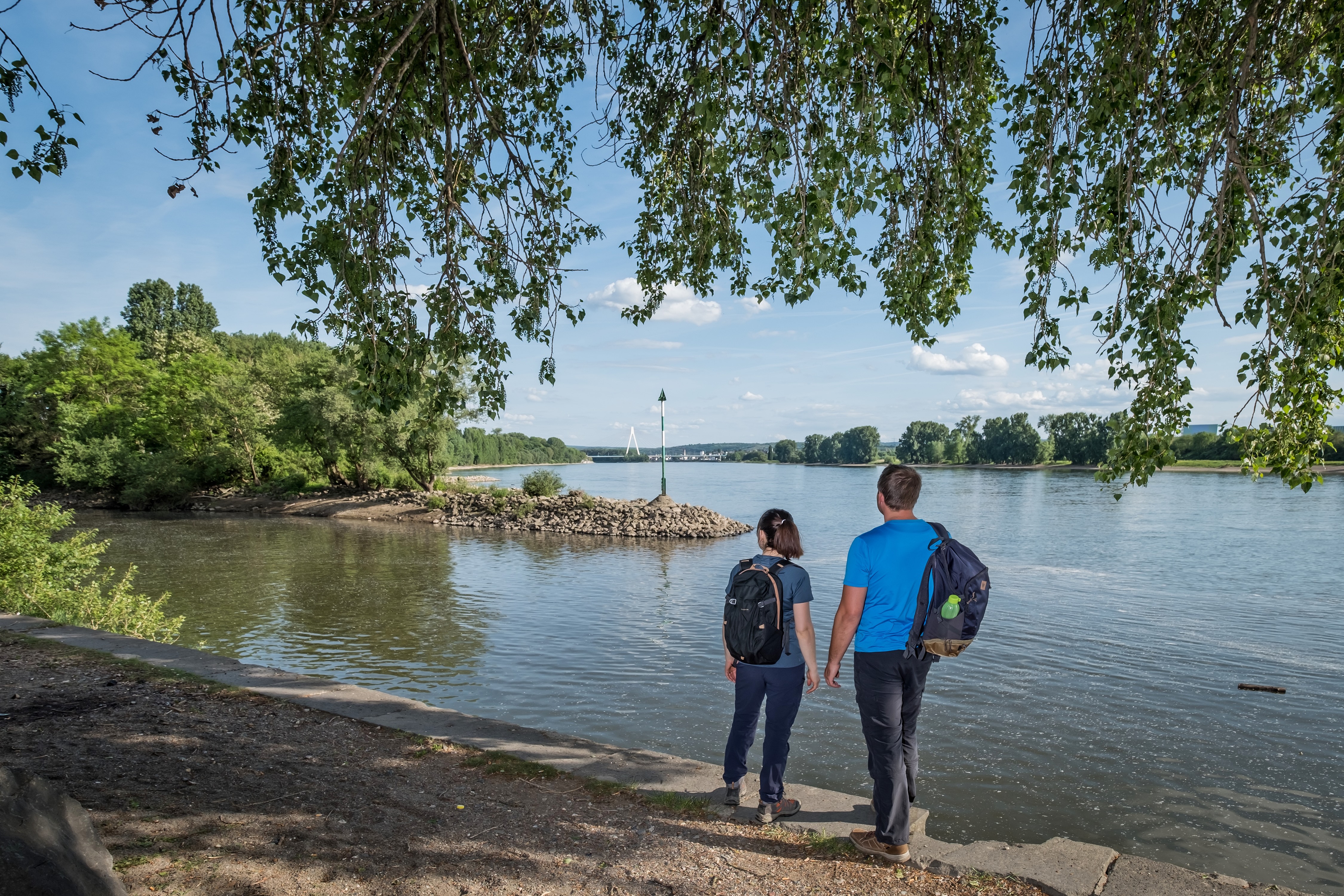 Zwei Personen halten Händchen am Ufer eines Flusses unter Bäumen