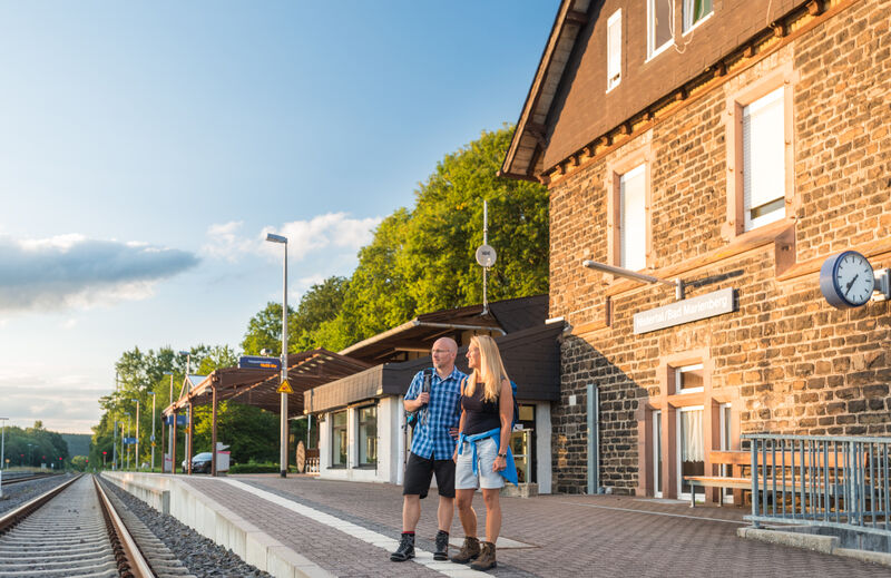 Wanderer stehen am Gleis eines Bahnhofs mit historischem Gebäude